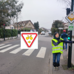 Au pied d'un feu rouge, une cycliste avec un gilet jaune indique avec son bras tendu la droite. Sur sa gauche un panneau M12 Droite