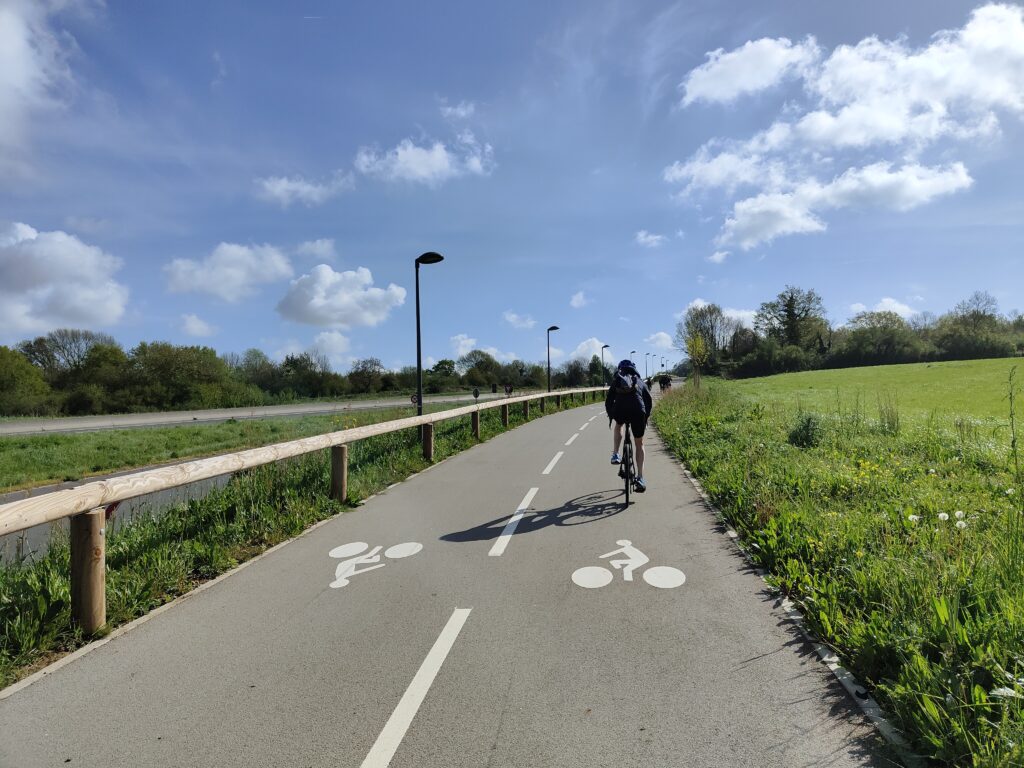 Cycliste de dos, avec un casque, sur une piste cyclable le long d'une route départementale hors agglomération, par beau temps
