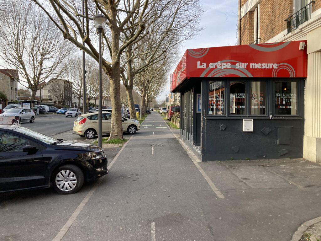 vue d'une piste cyclable, avec une voiture stationnée à sa limite sur la gauche, et une extension de restaurant sur sa droite.